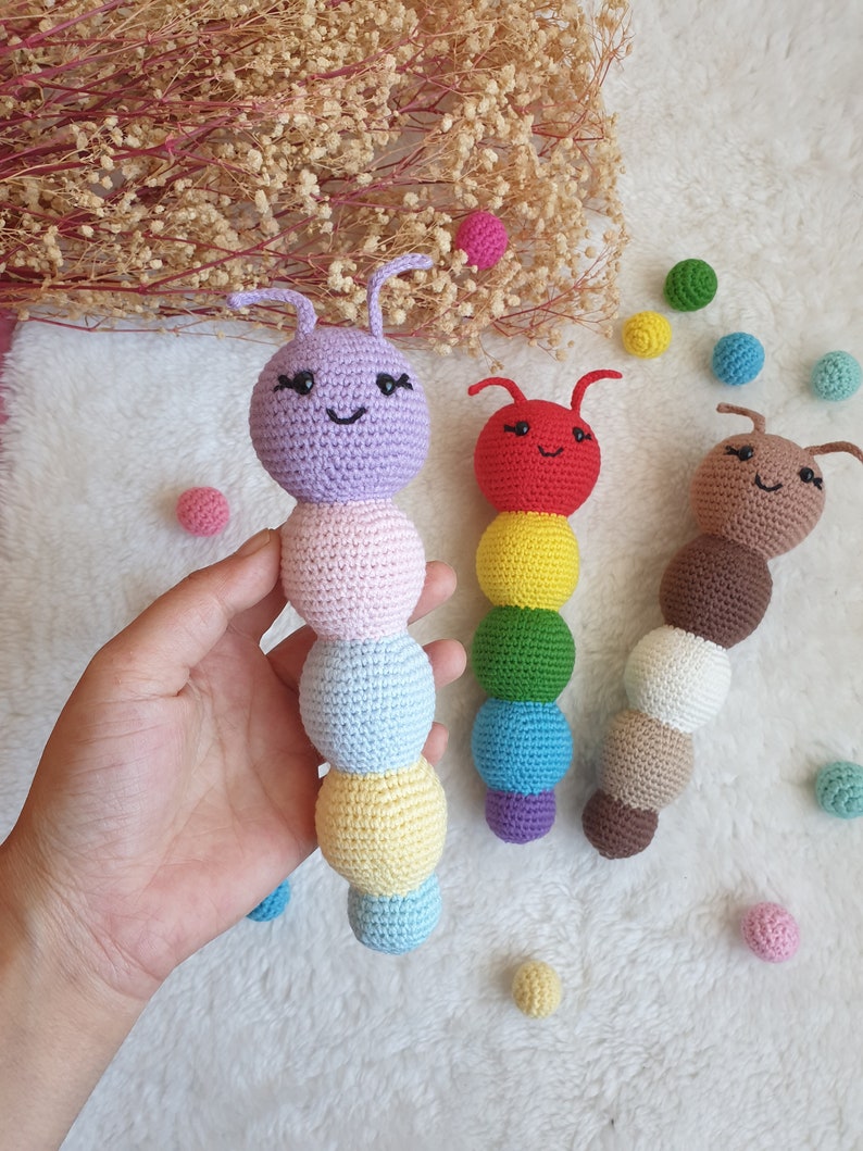 May include: Three crocheted rainbow-coloured caterpillars with smiling faces. The caterpillars are made of different coloured yarn, including purple, pink, blue, yellow, green, red, brown, and white.