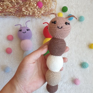 May include: A brown crocheted caterpillar toy with a smiling face and black eyes. The caterpillar is made of multiple crocheted balls in different colours, including brown, white, and beige. The toy is held in a hand against a white background.
