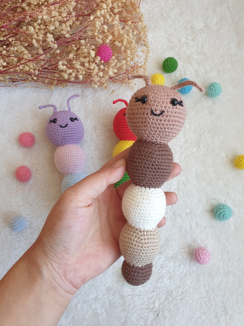 May include: A brown crocheted caterpillar toy with a smiling face and black eyes. The caterpillar is made of multiple crocheted balls in different colours, including brown, white, and beige. The toy is held in a hand against a white background.