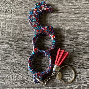 Könnte beinhalten: Ein Schlüsselanhänger aus rotem, weißem und blauem Glitzer mit einem Quastenanhänger. Der Schlüsselanhänger hat eine Schlaufe zum Anbringen an einer Tasche oder Schlüssel.