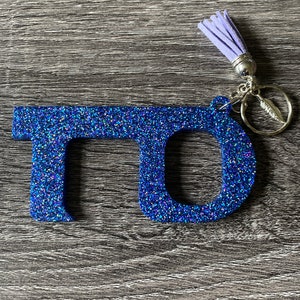 Könnte beinhalten: Ein blauer Glitzer-Türöffner-Schlüsselanhänger mit einem lilafarbenen Quasten. Der Schlüsselanhänger ist so konzipiert, dass er hilft, das Berühren von Oberflächen in der Öffentlichkeit zu vermeiden.