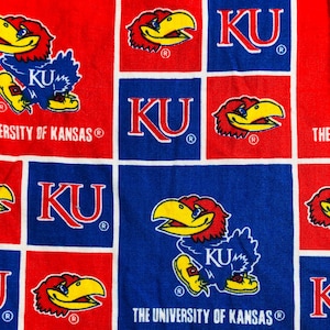 May include: A red and blue patterned fabric featuring the University of Kansas Jayhawk mascot and "KU" lettering. The design is a repeating grid of squares with the university's logo and text.