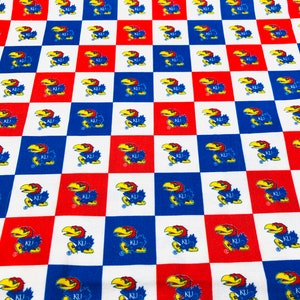 May include: Fabric with a repeating pattern of the University of Kansas Jayhawk mascot in a checkerboard design. The squares alternate between red, white, and blue, with the Jayhawk logo in each square. The fabric is likely cotton or a similar material.