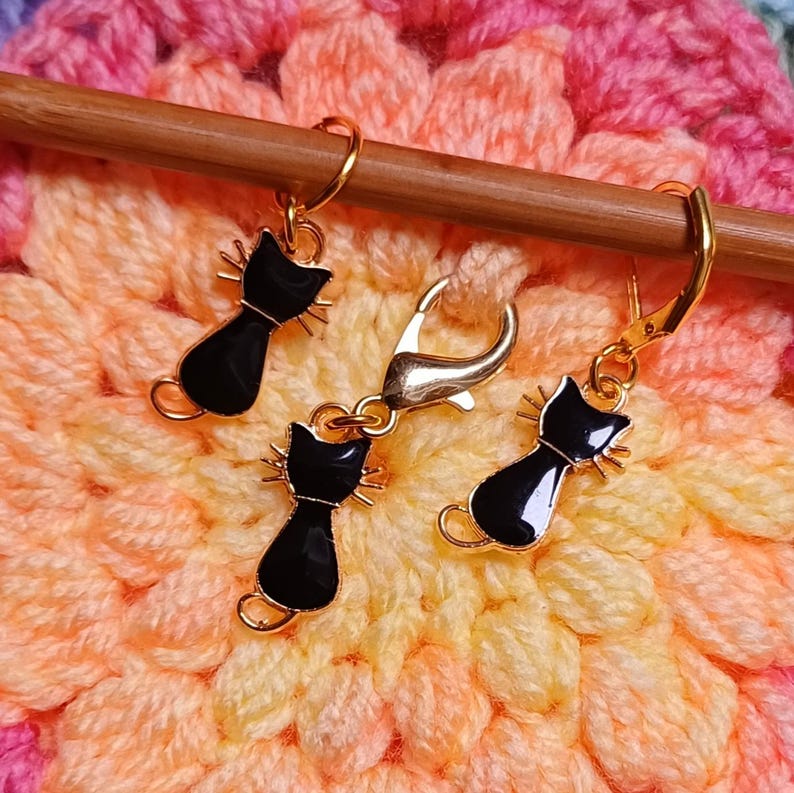 Black cat themed stitch marker with gold clasps and black and gold enamel cat charms, displayed on a wooden knitting needle over colourful crochet fabric—cute and functional stitch marker accessories for knitting and crochet projects. Three stitch markers displayed to show the three clasp options - lobster clasp, lever back and O Ring. Listing is for one single stitch marker.