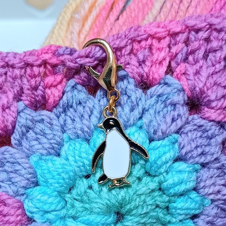 May include: A gold-colored stitch marker with a penguin charm. The penguin is black and white with a white belly and black wings and feet. The marker is attached to a crocheted blanket with pink, purple, and blue yarn.