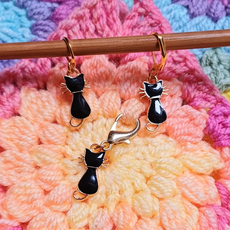 Black cat themed stitch marker with gold clasps and black and gold enamel cat charms, displayed on a wooden knitting needle over colourful crochet fabric—cute and functional stitch marker accessories for knitting and crochet projects. Three stitch markers displayed to show the three clasp options - lobster clasp, lever back and O Ring. Listing is for one single stitch marker.