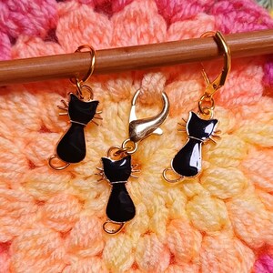 Black cat themed stitch marker with gold clasps and black and gold enamel cat charms, displayed on a wooden knitting needle over colourful crochet fabric—cute and functional stitch marker accessories for knitting and crochet projects. Three stitch markers displayed to show the three clasp options - lobster clasp, lever back and O Ring. Listing is for one single stitch marker.