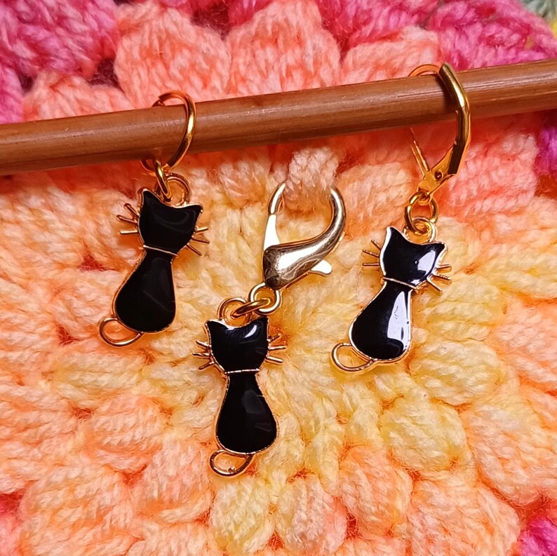 Black cat themed stitch marker with gold clasps and black and gold enamel cat charms, displayed on a wooden knitting needle over colourful crochet fabric—cute and functional stitch marker accessories for knitting and crochet projects. Three stitch markers displayed to show the three clasp options - lobster clasp, lever back and O Ring. Listing is for one single stitch marker.