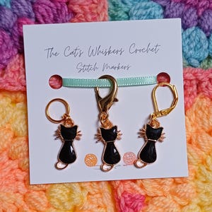 Black cat themed stitch marker with gold clasps and black and gold enamel cat charms, displayed on a white card with green ribbon over colourful crochet fabric—cute and functional stitch marker accessories for knitting and crochet projects. Three stitch markers displayed to show the three clasp options - lobster clasp, lever back and O Ring. Listing is for one single stitch marker.