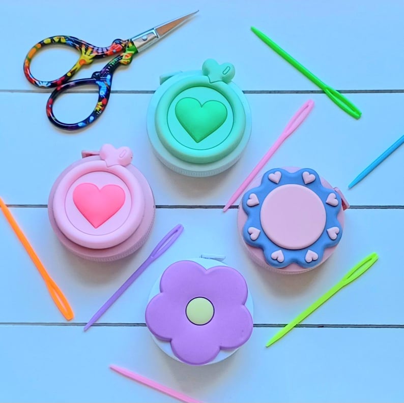 May include: Four colorful round containers with different designs: a green heart, a pink heart, a pink flower, and a blue flower. Each container has a small loop on top and is surrounded by colorful needles.