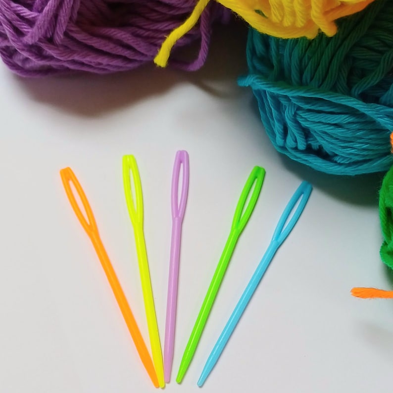 K&ouml;nnte beinhalten: F&uuml;nf bunte Plastik-Garnnadeln in Orange, Gelb, Hellgr&uuml;n, Hellblau und Lila auf einer wei&szlig;en Oberfl&auml;che.