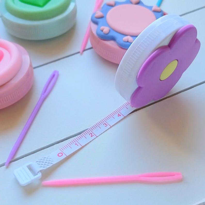 May include: A white and purple flower-shaped tape measure with a white retractable tape marked with numbers 0 through 4. The tape measure is on a white surface with pink and purple thread.