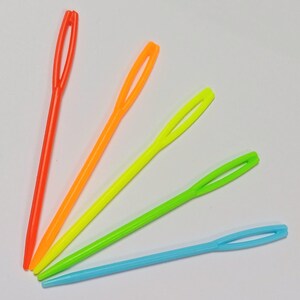 K&ouml;nnte beinhalten: F&uuml;nf Plastik-Garnnadeln in verschiedenen leuchtenden Farben: Orange, Gelb, Gr&uuml;n, Limettengr&uuml;n und Blau.