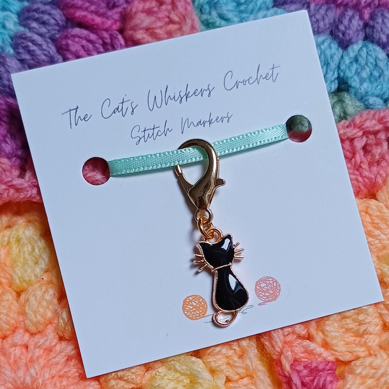Black cat themed stitch marker with gold clasp and black and gold enamel cat charm, displayed on a white card with green ribbon over colourful crochet fabric—cute and functional stitch marker accessories for knitting and crochet projects.