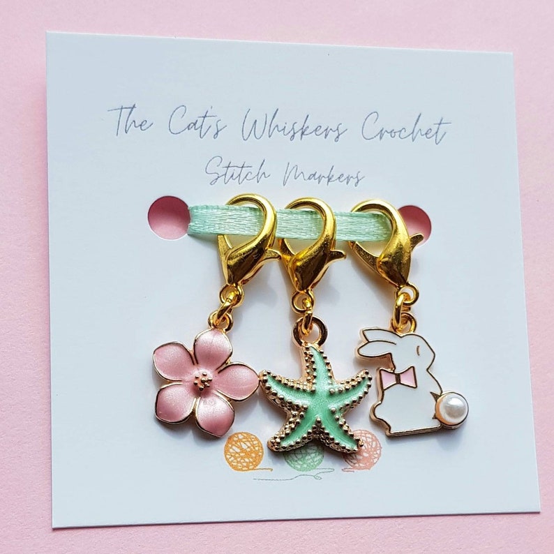 May include: Three gold stitch markers with a pink flower, a green starfish, and a white bunny charm. The charms are attached to the markers with a gold loop. The markers are displayed on a white card with the text "The Cat's Whiskers Crochet Stitch Markers".