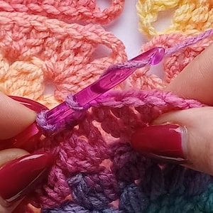 Pu&ograve; includere: Primo piano di una persona che lavora a maglia con un uncinetto viola. L'uncinetto viene utilizzato per tirare un'asola di filo attraverso un'altra asola di filo. Il filo &egrave; di vari colori, tra cui rosa, arancione, giallo e verde.