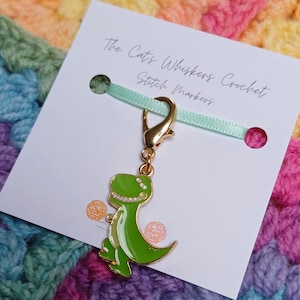May include: A green dinosaur stitch marker with gold-colored hardware. The marker is attached to a white card with the text "The Cat's Whiskers Crochet Stitch Markers" and a light green ribbon. The background is a colorful, textured fabric.