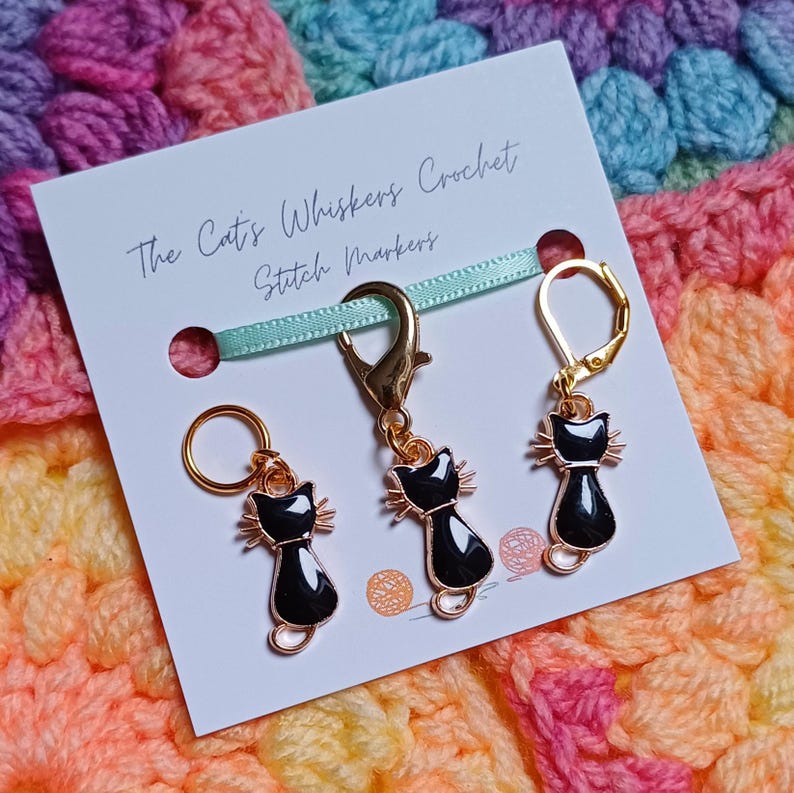 Black cat themed stitch marker with gold clasps and black and gold enamel cat charms, displayed on a white card with green ribbon over colourful crochet fabric—cute and functional stitch marker accessories for knitting and crochet projects. Three stitch markers displayed to show the three clasp options - lobster clasp, lever back and O Ring. Listing is for one single stitch marker.