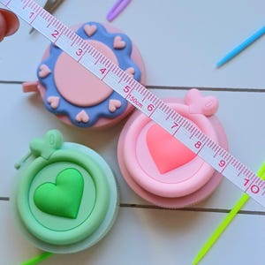 May include: Three colorful tape measures with heart designs. The tape measures are pink, green, and blue. Each tape measure has a different heart design. The tape measures are on a white surface.