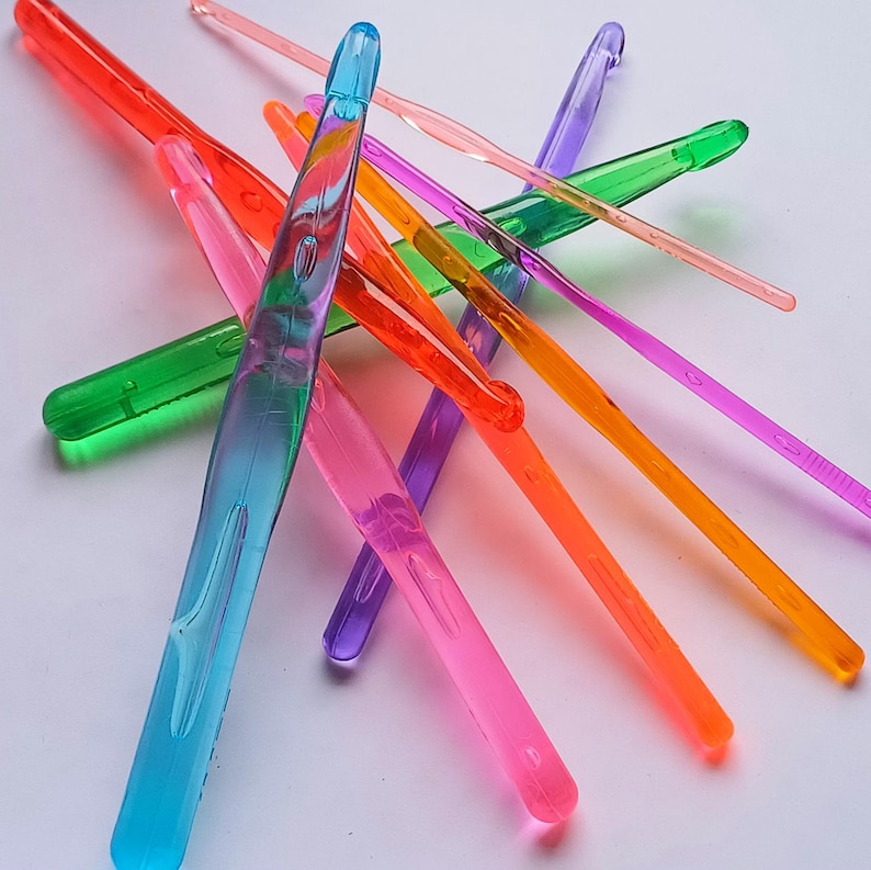 Pu&ograve; includere: Un set di uncinetti in plastica colorati di diverse dimensioni. Gli uncinetti sono disposti a croce su una superficie bianca.