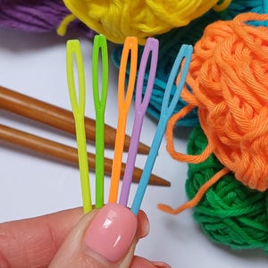 K&ouml;nnte beinhalten: F&uuml;nf bunte Plastik-Garnnadeln, mit einer gr&uuml;nen, orangefarbenen, violetten, blauen und gelben Nadel, werden in einer Hand vor einem Hintergrund aus buntem Garn gehalten.