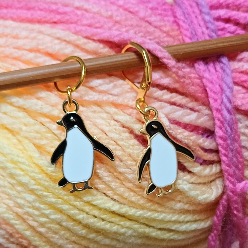 May include: Two gold penguin stitch markers. One marker is a simple ring, the other has a hook closure. Both penguins are black and white with gold accents.