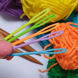 K&ouml;nnte beinhalten: Vier bunte Plastik-Garnnadeln, mit einer gr&uuml;nen, orangefarbenen, violetten und blauen Nadel, liegen auf Garnkn&auml;ueln in verschiedenen Farben, darunter lila, gelb, orange und gr&uuml;n.