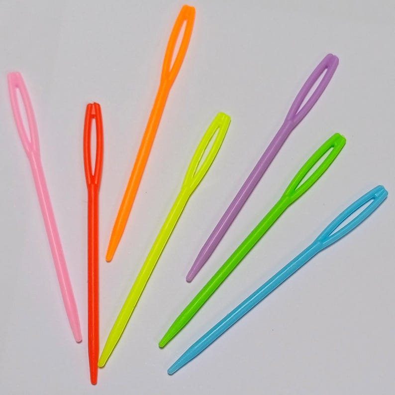 K&ouml;nnte beinhalten: Ein Satz mit sieben bunten Plastiknadeln zum Einf&auml;deln von Garn. Die Nadeln sind in den Farben Pink, Orange, Gelb, Gr&uuml;n, Hellblau und Lila.