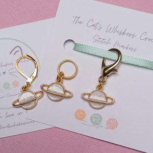 Pode incluir: Três marcadores de ponto de ouro com pingentes de planetas brancos. Os marcadores são em forma de anel, fecho de lagosta e gancho. O texto "The Cat's Whiskers Crochet Stitch Markers" é visível no fundo.