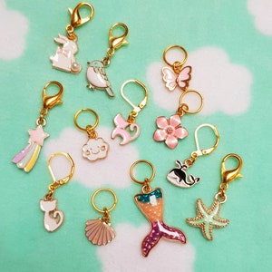 May include: A set of 12 colorful stitch markers with various charms, including a mermaid, a cat, a cloud, a star, a butterfly, a whale, a seashell, a flower, a bird, a rabbit, and a starfish. The stitch markers are made of metal and have a gold finish.