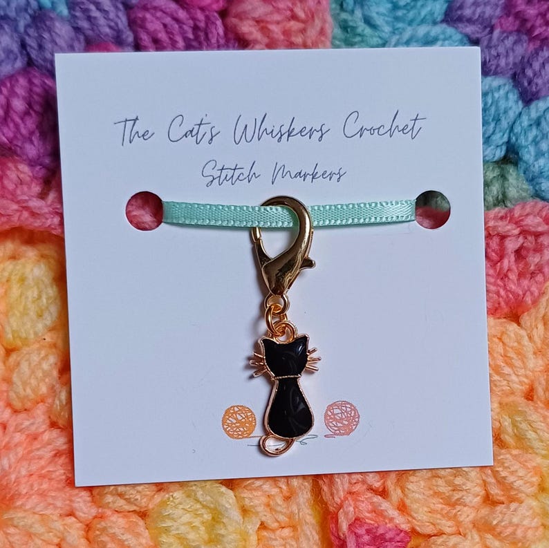 Black cat themed stitch marker with gold clasp and black and gold enamel cat charm, displayed on a white card with green ribbon over colourful crochet fabric—cute and functional stitch marker accessories for knitting and crochet projects.