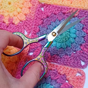 May include: Rainbow-hued embroidery scissors with a floral pattern, positioned above a vibrant crocheted square. The scissors feature sharp blades and ornate handles, ideal for various craft activities.
