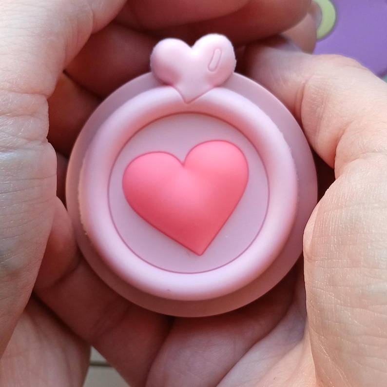 May include: A pink silicone phone ring with a heart-shaped design. The ring has a raised heart in the center and a smaller heart on top.
