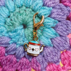 May include: A gold-colored charm with a small white teapot design, featuring a red and blue lid, and black spade, diamond, and pink heart details. The charm is attached to a gold clasp, set against a colorful, crocheted background.
