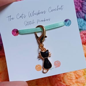 Black cat themed stitch marker with gold clasp and black and gold enamel cat charm, displayed on a white card with green ribbon over colourful crochet fabric—cute and functional stitch marker accessories for knitting and crochet projects.