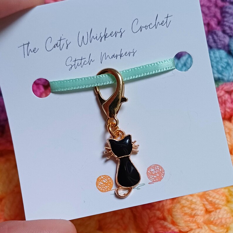 Black cat themed stitch marker with gold clasp and black and gold enamel cat charm, displayed on a white card with green ribbon over colourful crochet fabric—cute and functional stitch marker accessories for knitting and crochet projects.