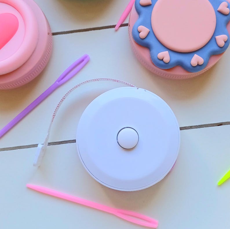 May include: A white retractable tape measure with a pink and blue decorative case. The case has a heart design.