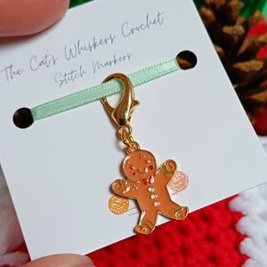 Op de afbeelding: Een goudkleurige gingerbread man steekmarkeerder met een rode strik en witte knopen. De bedel is bevestigd aan een gouden sluiting en een lichtgroen lint. De kaart leest "The Cat's Whiskers Crochet Stitch Markers."