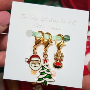 May include: Three gold stitch markers with Christmas themed charms. The charms are a Santa Claus, a Christmas tree, and a reindeer. The text "The Cat's Whiskers Crochet Stitch Markers" is printed on the white card.