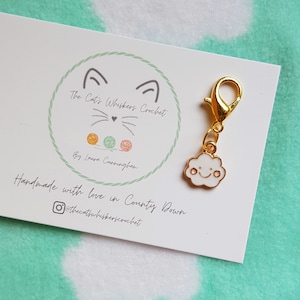 May include: A gold-colored stitch marker with a white cloud charm. The cloud has a smiling face with two small eyes and a mouth. The stitch marker is attached to a card with the text "The Cat's Whiskers Crochet" and "Handmade with love in County Down".