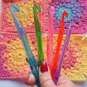 May include: A set of five colourful plastic crochet hooks in various sizes. The hooks are held in a hand and are displayed against a background of crocheted squares.