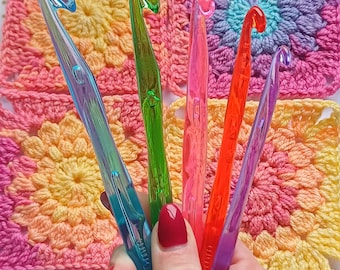 May include: A set of five colourful plastic crochet hooks in various sizes. The hooks are held in a hand and are displayed against a background of crocheted squares.