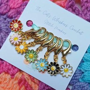 May include: A collection of gold-tone stitch markers, each adorned with a colourful daisy charm. The daisy charms feature shades of red, pink, yellow, green, blue, black, and white. The markers are displayed on a card with the text "The Cats Whiskers Crochet Stitch Markers".