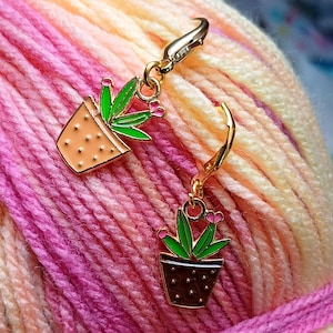 May include: Two gold-tone metal stitch markers shaped like potted plants with green leaves and pink flowers. The markers are on a background of pink and yellow yarn.