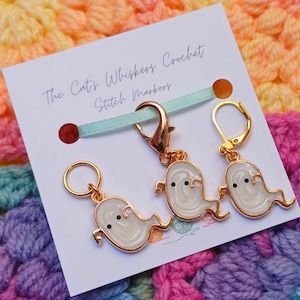 Op de afbeelding: Een set van drie goudkleurige stekenmarkeerders in de vorm van geesten, met witte emaille en zwarte stipogen. De markeerders zijn bevestigd aan een kaart met de tekst "The Cat's Whiskers Crochet Stitch Markers".