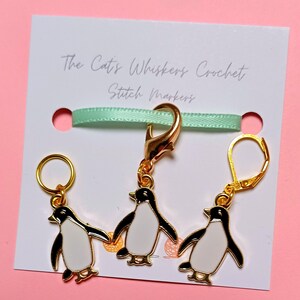 May include: Three gold penguin stitch markers. One marker is a clip, and the other two are loop markers. The markers are on a white card with the text "The Cat's Whiskers Crochet Stitch Markers".