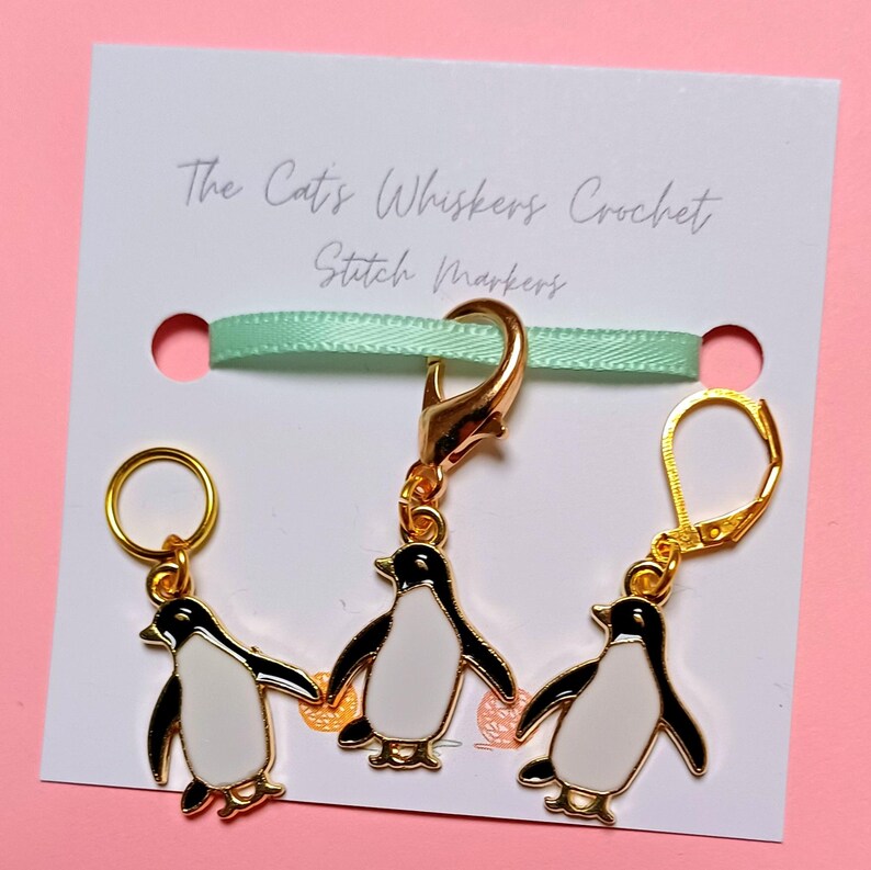 May include: Three gold penguin stitch markers. One marker is a clip, and the other two are loop markers. The markers are on a white card with the text "The Cat's Whiskers Crochet Stitch Markers".