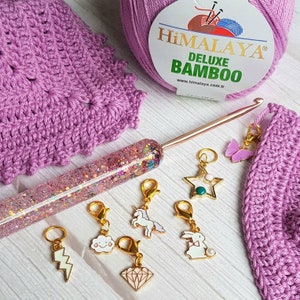 May include: A close-up of a set of colorful stitch markers with various designs, including a unicorn, a cloud, a lightning bolt, a diamond, a star, a rabbit, and a butterfly. The stitch markers are on a white surface with a pink crochet project and a crochet hook.