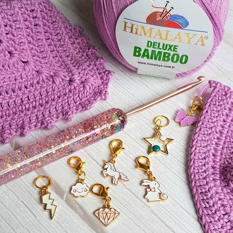 May include: A close-up of a set of colorful stitch markers with various designs, including a unicorn, a cloud, a lightning bolt, a diamond, a star, a rabbit, and a butterfly. The stitch markers are on a white surface with a pink crochet project and a crochet hook.