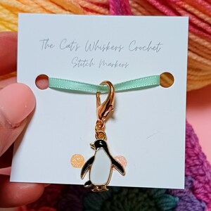 May include: Gold penguin stitch marker with a black and white design. The marker is attached to a gold clasp and a light green ribbon. The card reads "The Cat's Whiskers Crochet Stitch Markers".